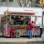 A bustling street vendor selling tacos in a colorful open-air stall on a sunny day.